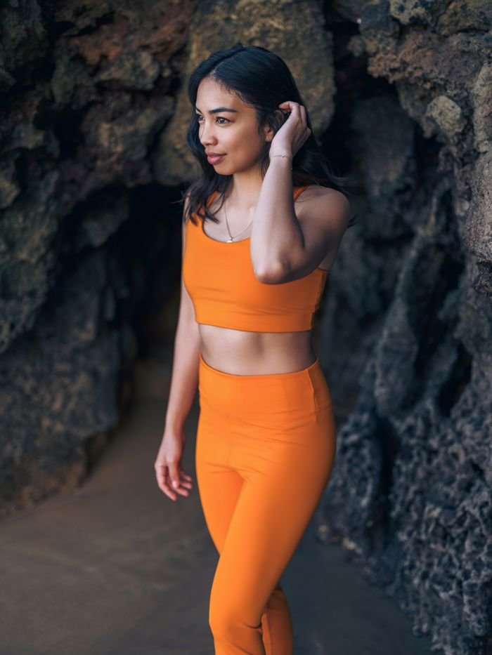 Brunette woman wearing orange activewear posing confidently near rock formations in Lisbon, Portugal.