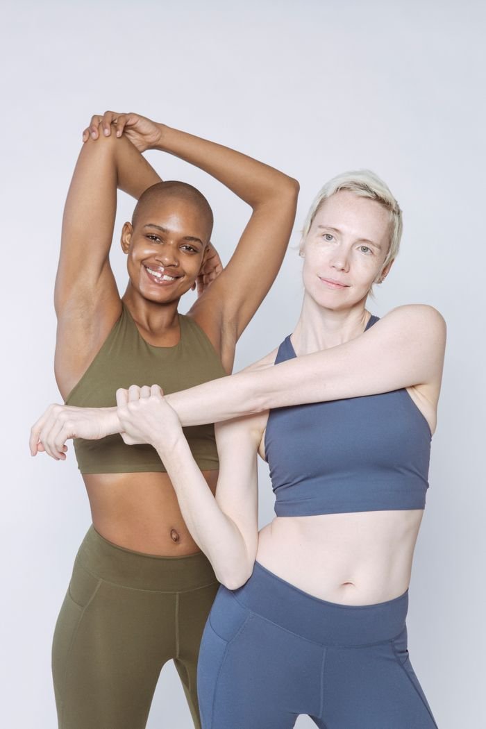 Positive diverse ladies in activewear stretching arms while standing in studio and looking at camera